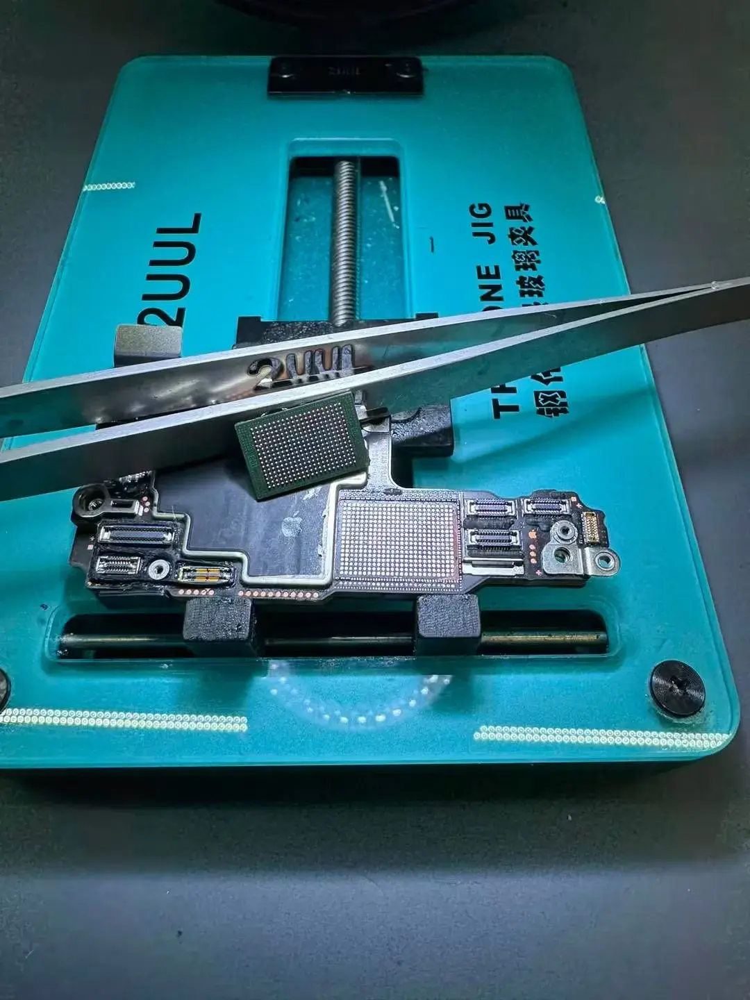 Tweezers lifting a chip off an iPhone logic board held in a repair fixture, with the solder ball grid exposed