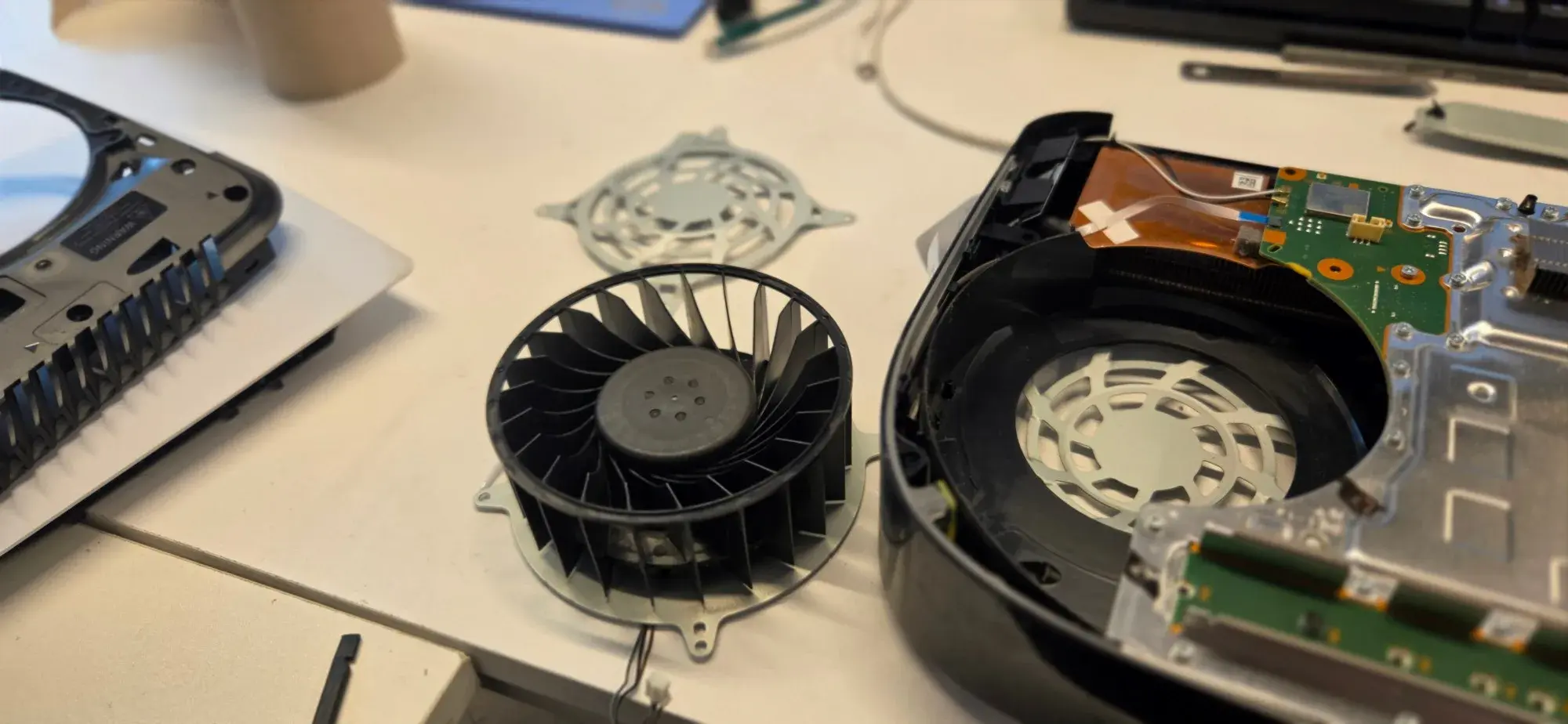 A PS5 blower fan removed and set on the repair bench beside the opened console and its fan guard