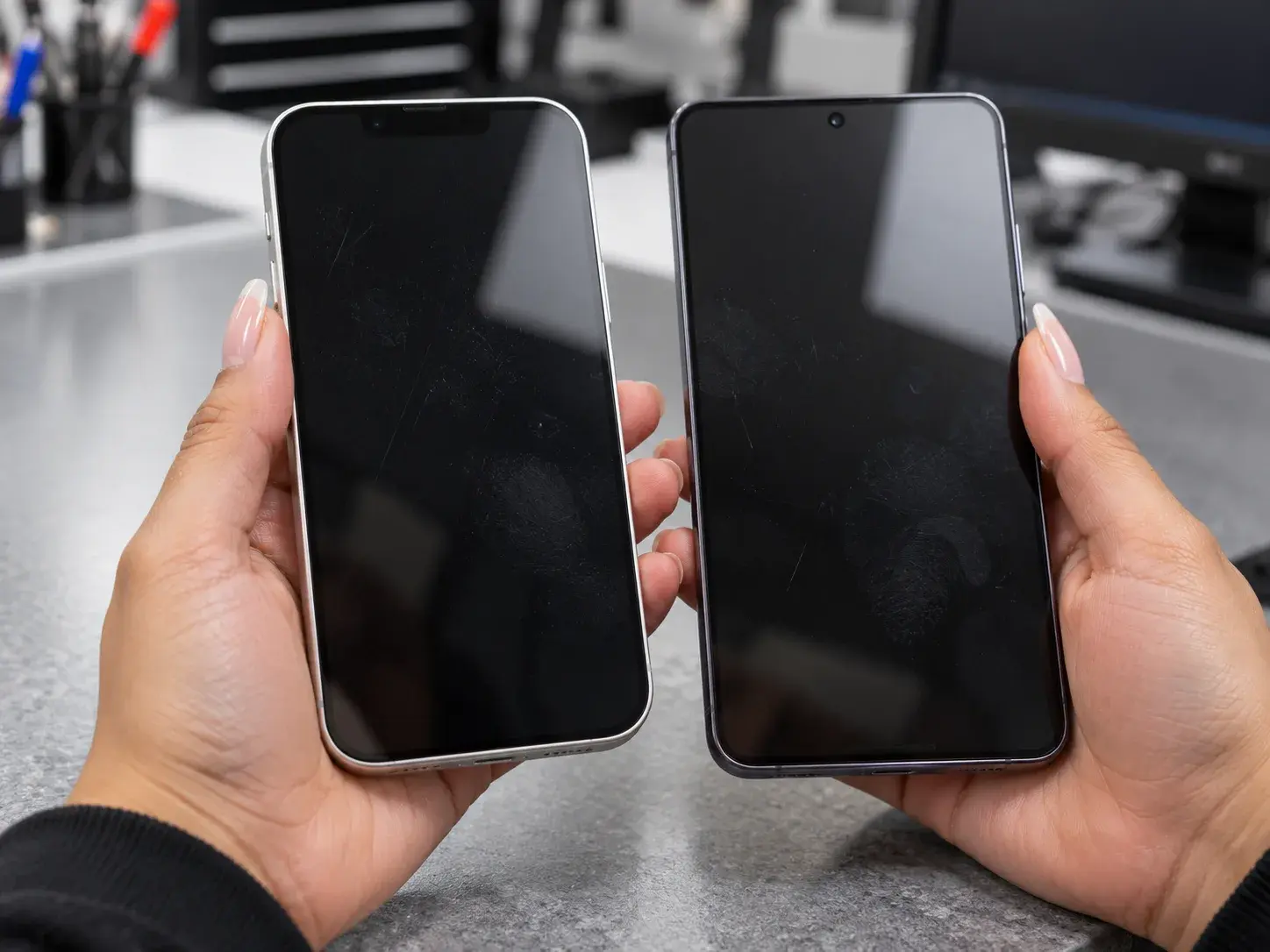Two hands holding an iPhone and a Samsung Galaxy side by side over a shop counter, both screens off